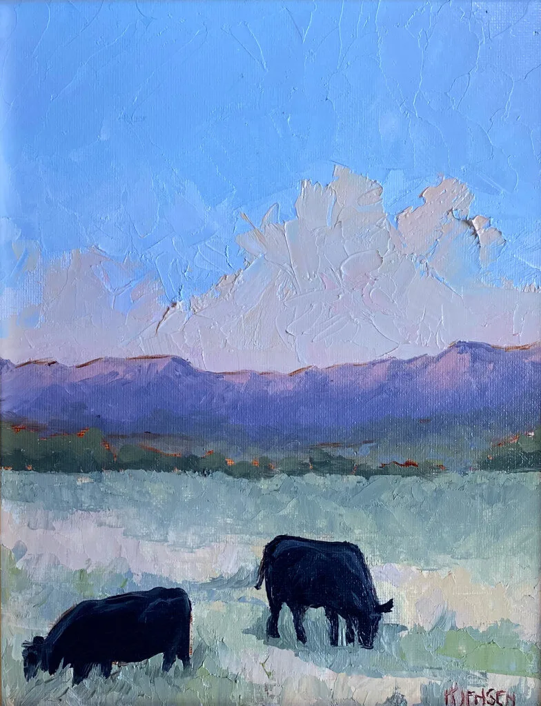 Two black cows grazing in pale field below purple mountains and tall white clouds