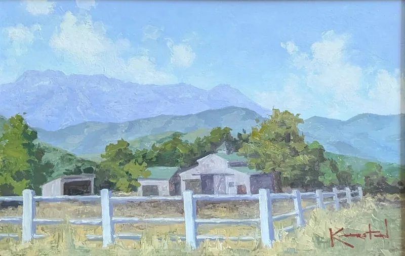 White barn and sheds behind a fence with green foothills and mountains rising behind.