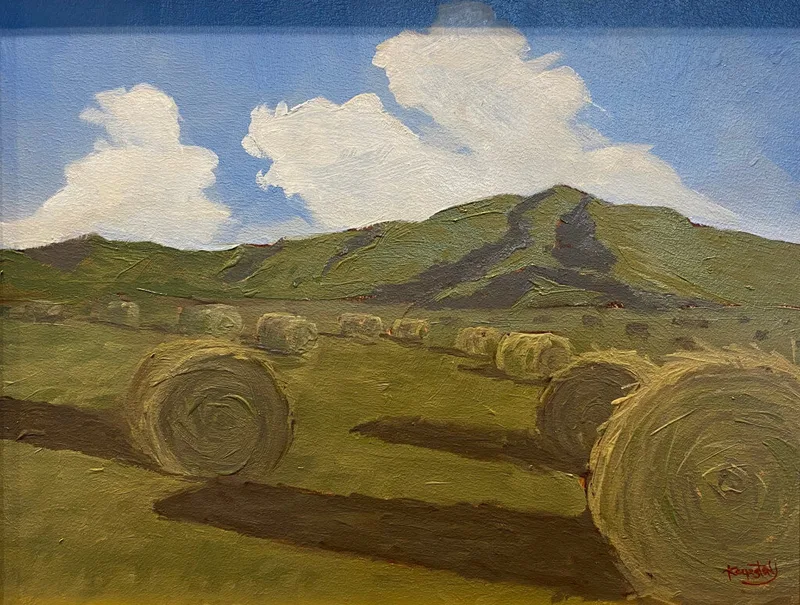 Round and rectangular hay bales scattered across green field beneath rolling hills and clouds