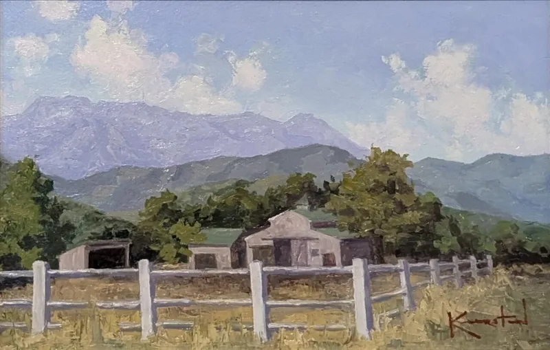 Rustic barn behind a white fence with rolling foothills and hazy mountains behind.